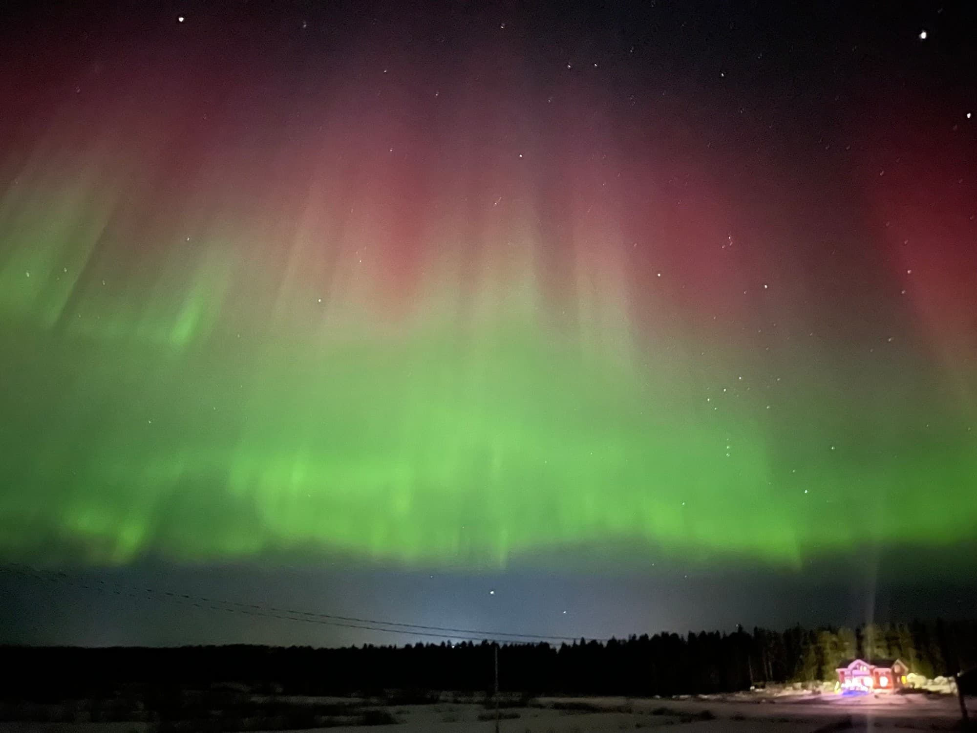 Green and red aurora over the snow, the foundry house lit at the tree line